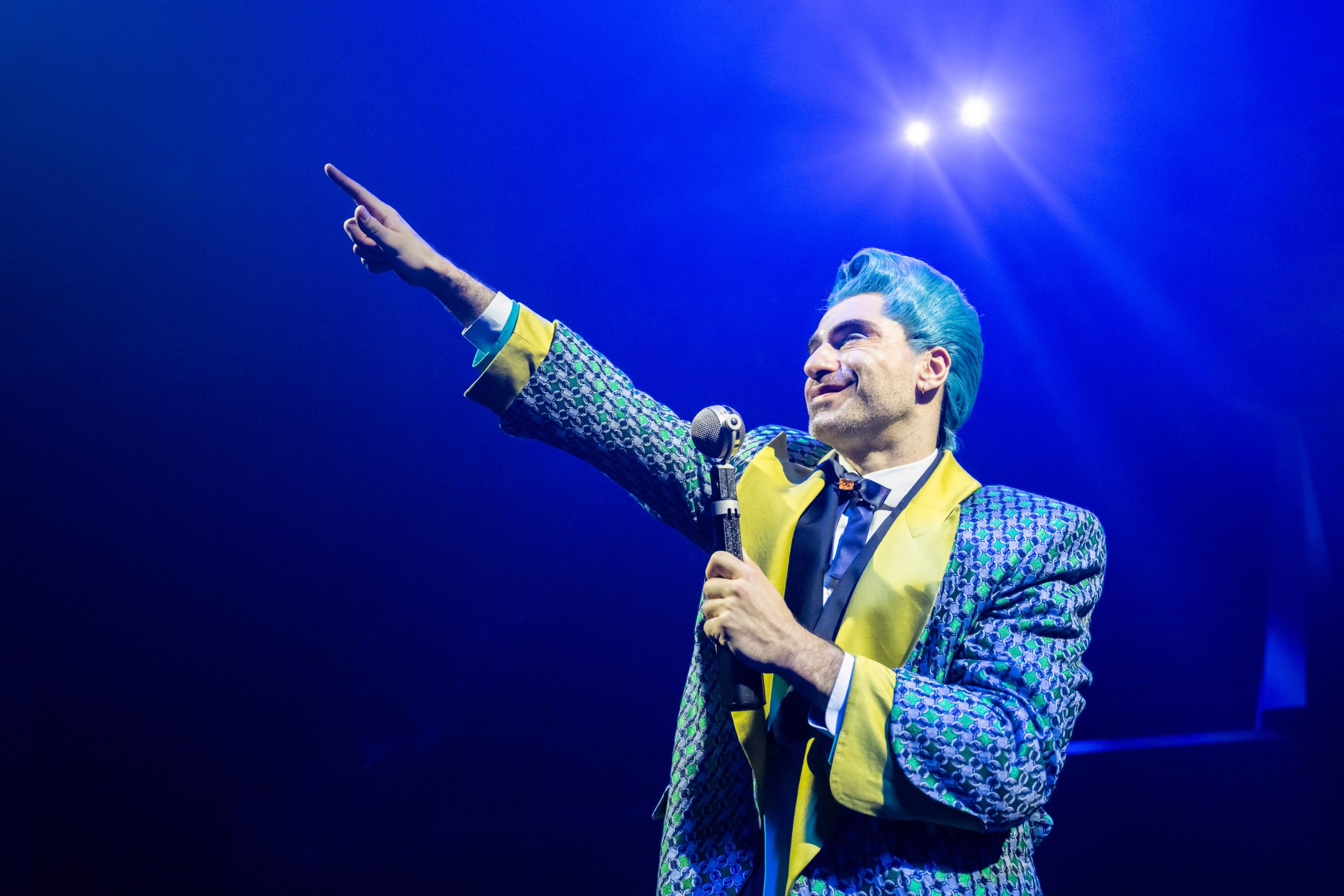 Caesar Flickman holding a mic in one hand and pointing upwards with the other in The Hunger Games: On Stage in London